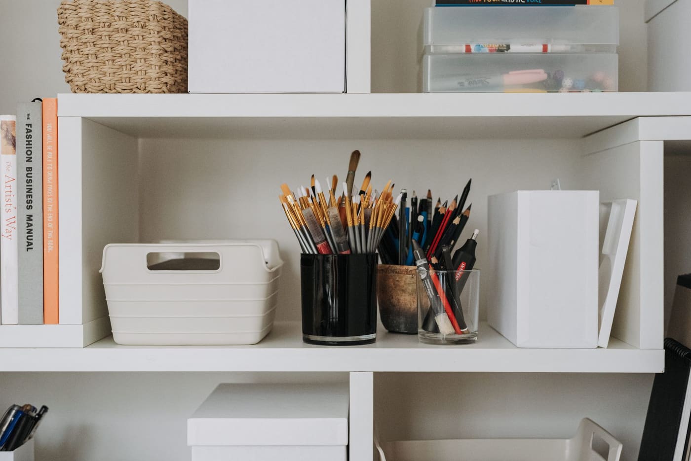 A white shelving unit holding lidded boxes, a cup of brushes, and an open storage bin