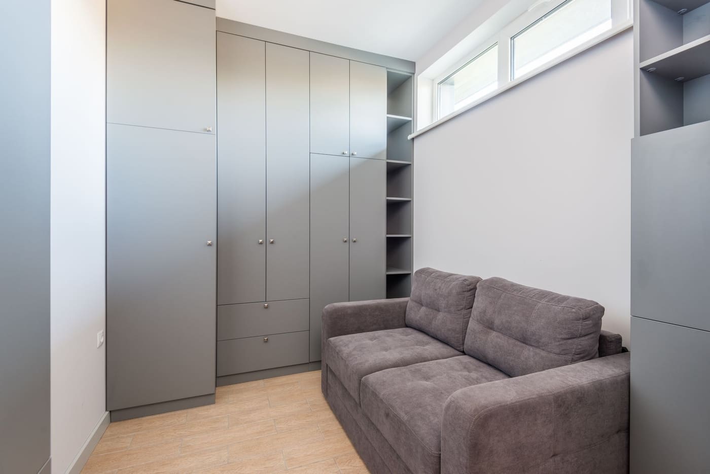 A compact gray sofa beside a built-in grey wardrobe in a small room with a high window