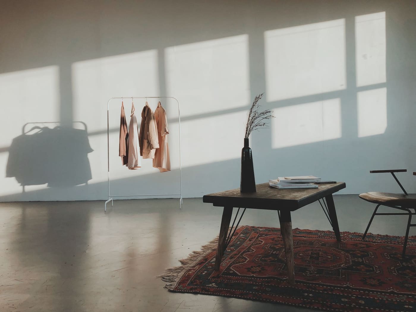 A sunlit room with pale walls, a rug, a small table, and a clothes rail by a window